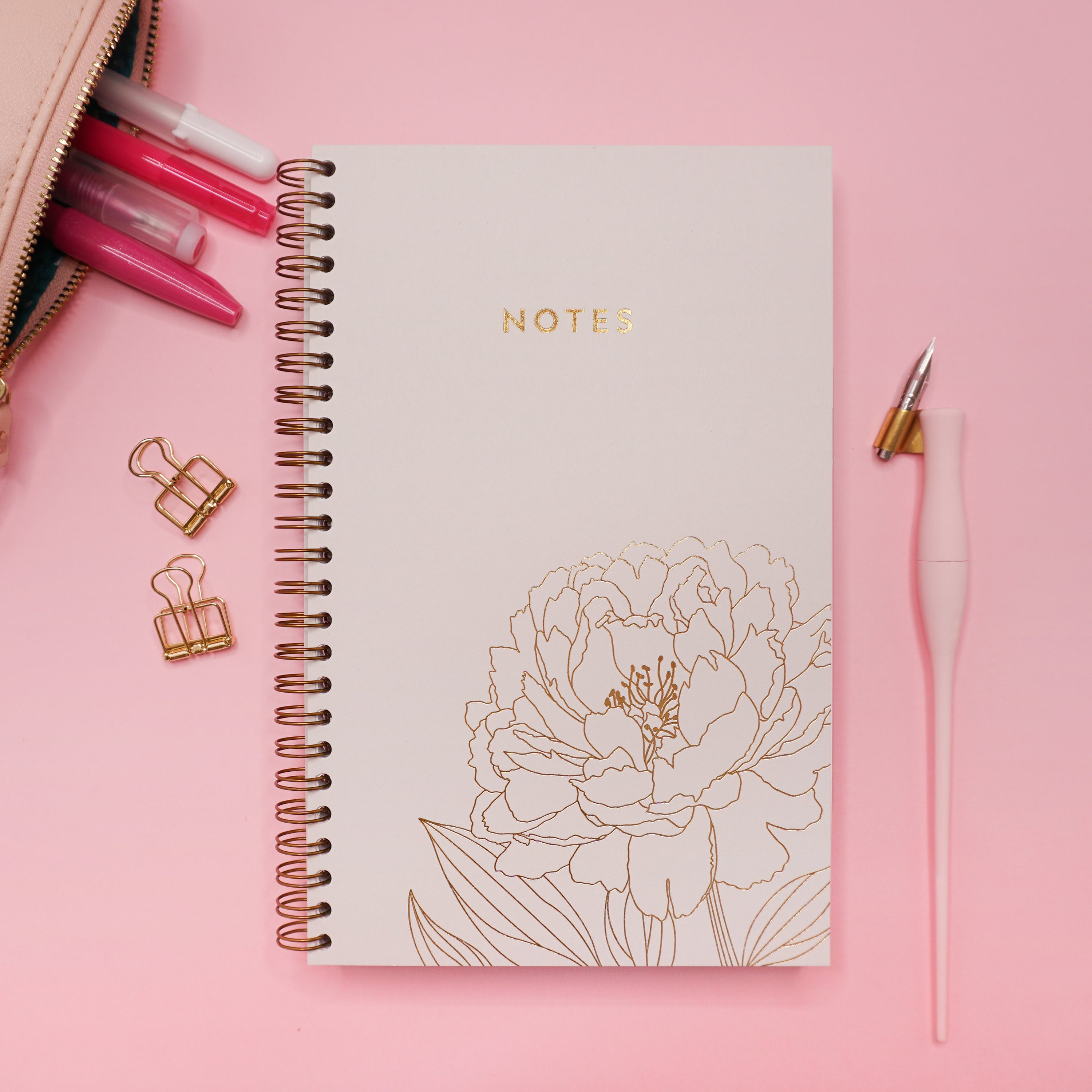 Le cahier pivoine, accompagné d'une trousse à crayon et une plume sur fond rose.